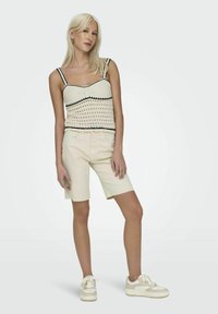 Top de ganchillo color crema con ribete negro, combinado con pantalones cortos beige claros y zapatillas beige. Los pantalones cortos presentan un dobladillo sin terminar.