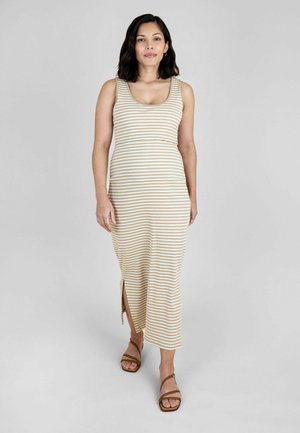 Femeie în picioare pe fundal gri, purtând o rochie midi fără mâneci cu dungi bej și albe, cu deschidere laterală, și sandale maro cu barete.