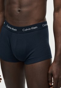 Κοντινό πλάνο ενός σκούρου δέρματος ανδρικού κορμού που φοράει σκούρα μπλε boxer Calvin Klein με λογότυπο στη μέση.