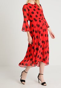 Robe rouge à pois avec manches évasées 3/4, tissu fluide, longueur genou, et sandales à talons noires. Texture lisse avec une coupe ample.
