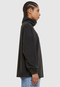 Pullover nero ampio con colletto alto, maniche lunghe e polsini a costine. Realizzato in un morbido tessuto texturizzato. Indossato con jeans azzurri chiari.