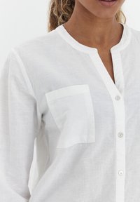 Hvid linned skjorte med knaplukning foran, rund hals og en enkelt brystlomme. Blød tekstur, let design og minimale detaljer.