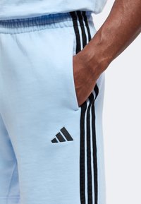 Χέρι στην τσέπη ανοιχτό μπλε φόρμα adidas με μαύρες ρίγες και λογότυπο στο πλάι.
