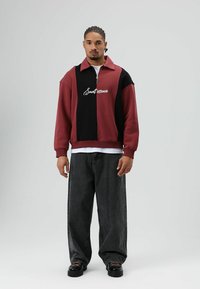 Sweat-shirt zippé bordeaux et noir avec col, arborant un lettrage de logo blanc et des poignets côtelés, assorti à un jean noir ample.