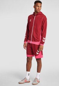 Felpa sportiva rossa con motivo a pois, accenti rosa, pantaloni corti abbinati con design a chevron bianco, e sneakers beige con accenti rossi.