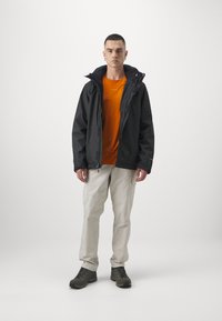Sort vandafvisende jakke med hætte, orange kortærmet skjorte, beige bukser og grønne vandrestøvler, står på en ensartet baggrund.