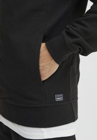 Svart strukturert sweatshirt med kengurulomme; har en hvit lagdelt kant og et merke på den nedre siden som indikerer merkevaren og kolleksjonen.
