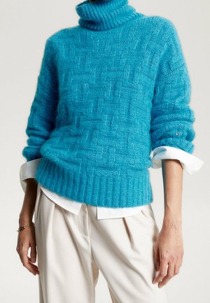 Türkiser Strickpullover mit hohem Rippkragen, strukturiertem Muster und umgeschlagenen Ärmeln, übereinander getragen mit einem weißen Hemd mit Kragen.