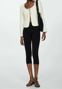 Veste ajustée en crème clair avec un décolleté en forme de scoop et deux fermoirs en argent, associée à des leggings noirs coupés et des sandales noires à lanières.