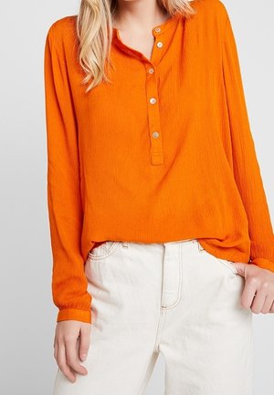 Kvinde iført en langærmet, knaplukket bluse i stærk orange, stoppet ned i højtailede, off-white bukser, med hånden hvilende i lommen.