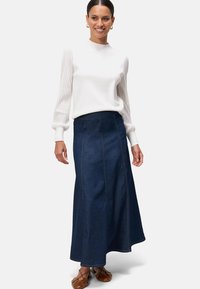 Jupe longue en denim foncé avec une taille ajustée et des détails de coutures verticales. Associée à un chemisier blanc avec des manches côtelées. Chaussures plates brunes.