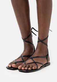 Tumman ihon sävyiset jalat, joilla on mustat litteät sandaalit, joissa on ohuet ristikkäiset hihnat ja nilkkaketju, valkoisella taustalla.