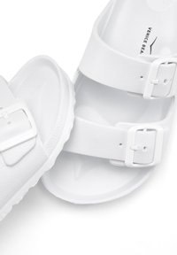 White sandals made of smooth synthetic material, featuring two adjustable straps with white buckles, textured outsole, and rounded shape.
