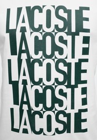 Επαναλαμβανόμενο πράσινο κείμενο "LACOSTE" τοποθετημένο σε εναλλασσόμενο μοτίβο πλέγματος σε λευκό φόντο.