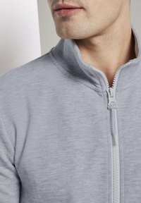 Chaqueta gris claro con cremallera y cuello alto, que presenta una tela texturizada y una cremallera blanca en contraste. Vista en primer plano del área del cuello.