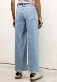 Jean large bleu clair avec une texture lisse, comprenant deux poches arrière et un onglet de taille beige. Associé à des baskets claires.