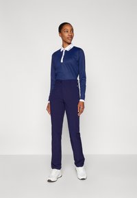 Navy modrá polokošile s dlouhým rukávem a bílým límcem, doplněná navy kalhotami. Model má na sobě bílé sportovní boty. Jednoduchý design, žádné vzory ani loga viditelná.