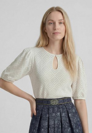 Vrouw met lang blond haar die een witte blouse met patroon draagt met pofmouwen en een sleutelgathals, gecombineerd met een marineblauwe bloemen geplooide rok.