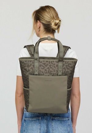 Woman with blonde hair in a bun wears a white shirt and blue jeans, carrying a gray leopard-print backpack with dual handles on her back.