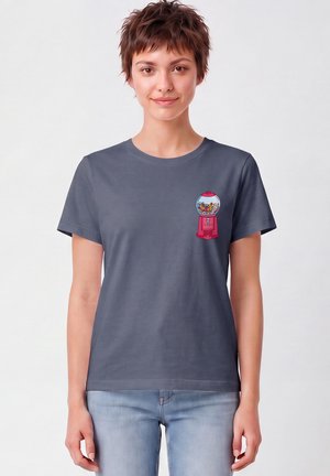 Jeune personne aux cheveux courts portant un t-shirt gris foncé avec un motif coloré de distributeur de chewing-gums sur le côté gauche de la poitrine, associé à un jean bleu clair.