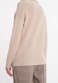 Beige ribbstickad tröja med krage, långa ärmar och lös passform. Bärs med bruna rutig byxor och visar texturerat tyg.