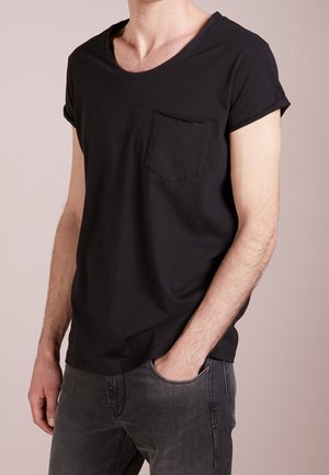 Homme portant un t-shirt noir uni à manches courtes avec une poche poitrine et un jean gris, une main dans la poche, fond neutre.