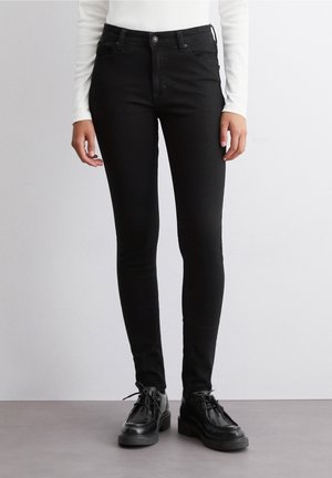 Persoon draagt zwarte skinny jeans, zwarte veterschoenen en een wit langermige top en staat op een grijze vloer.