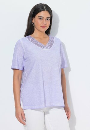 Frau mit langen dunklen Haaren, die ein lavendelfarbenes V-Ausschnitt-Shirt mit Spitzenbesatz und kurzen Ärmeln sowie weiße Hosen trägt, vor einem einfarbigen Hintergrund.