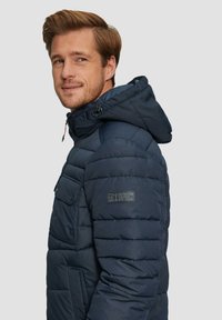 Marineblauwe gewatteerde jas met capuchon, voorzien van horizontale stiksels, voorkeurszakken en een logo-opdruk op de mouw. Glad nylonmateriaal.