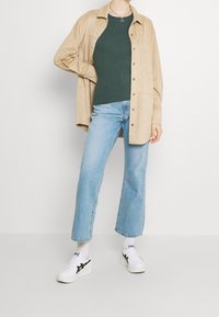 Beige mokkanahka-paita takki tummanvihreän ribbipaidan päällä, yhdistettynä vaaleansinisiin leveälahkeisiin farkkuihin ja valkoisiin lenkkareihin mustilla yksityiskohdilla.