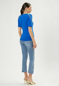 Blauw top met korte mouwen en schouderaccenten, gecombineerd met lichtblauwe cropped jeans. Model draagt gouden open teen hakken. Gladde stofstructuur.