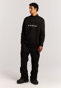 Schwarzes Langarmshirt mit "PROTEST"-Logo über der Brust, kombiniert mit schwarzen Hosen und schwarzen Stiefeln. Das Shirt hat eine lockere Passform und eine glatte Textur.