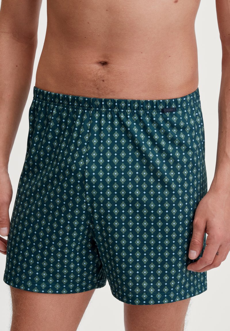 Shorts en coton vert foncé, présentant un motif en losanges vert clair et bleu. Ceinture élastique avec étiquette de marque sur le côté.