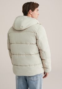 Chaqueta acolchada color beige claro con capucha, que presenta un diseño acolchado, costuras en los hombros y una textura suave y mate.