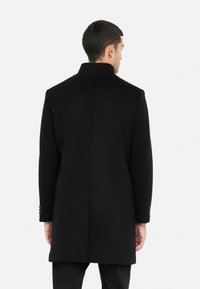 Cappotto nero in lana dal taglio dritto, collo alto e maniche lunghe. Presenta polsini con bottoni e una sola spaccatura sul retro.