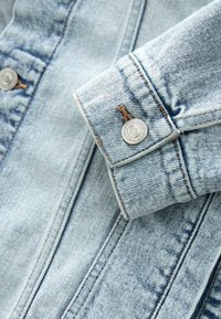 Lichtblauw denimjack met een manchet met een zilveren knoop, met stikseldetails en een gestructureerd oppervlak. Zichtbare nerf en ongelijke kleurvariaties.