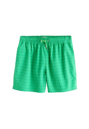 Joven de pie descalzo al aire libre con pantalones cortos verdes de baño con cordón, bajo un cielo azul claro junto a una pared blanca texturizada.