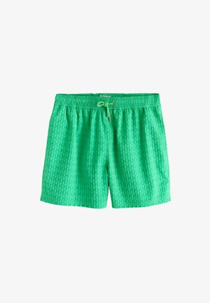 Ung mand står barfodet udendørs iført grønne badeshorts med snøre, under en klar blå himmel ved en tekstureret hvid væg.