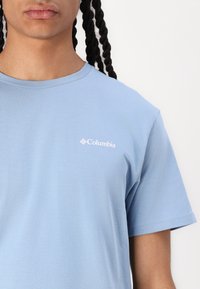 Mėlynos spalvos trumpomis rankovėmis marškinėliai, pagaminti iš švelnaus medvilnės, su balta "Columbia" logotipu ant kairės krūtinės. Lygi tekstūra, standartinio kirpimo.