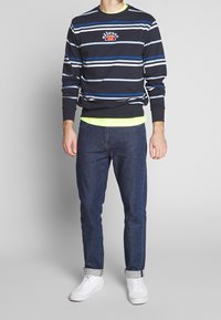 Uomo in camicia a righe blu navy a maniche lunghe, maglietta gialla fluo sotto, jeans scuri arrotolati e sneakers bianche, in piedi contro uno sfondo semplice.