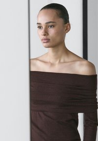 Brown off-the-shoulder knit top, featuring textured fabric with gathered detail. Smooth skin tone and minimalist background.
