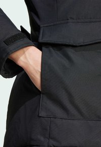 Black outerwear detail featuring a pocket, textured fabric, long sleeves, and a Velcro closure at the cuff.