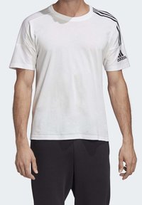 Bílá tričko s krátkým rukávem a černým logem adidas. Obsahuje tři černé pruhy na ramenou, zaoblený výstřih a jemnou texturu látky.