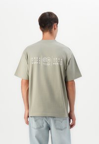 Mand i lysegrøn kreativ arbejds-T-shirt og lyseblå jeans, der står med ryggen til foran en ensfarvet hvid baggrund.