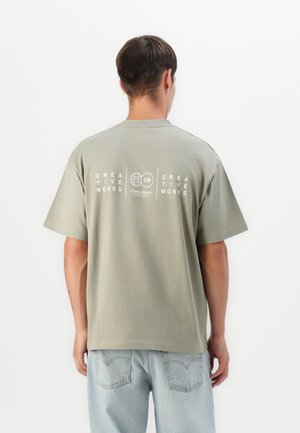 Man in light green creative works T-shirt and light blue jeans, facing away against a plain white background.