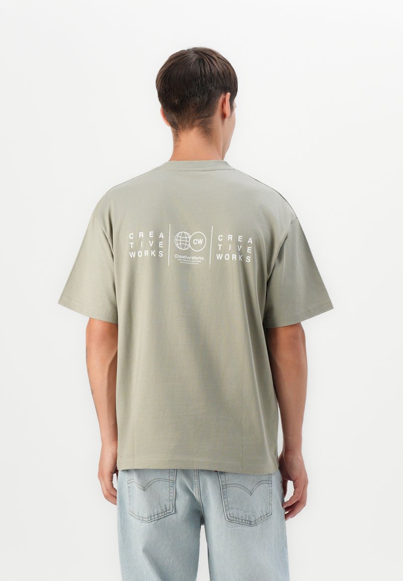 Mand i lysegrøn kreativ arbejds-T-shirt og lyseblå jeans, der står med ryggen til foran en ensfarvet hvid baggrund.