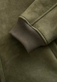 Olivenfarvet jakke med en glat faux suede tekstur og ribbede manchetter i en mørkere grøn nuance. Jakken har et strømlinet design.