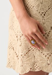 Yellow resin ring with an octagonal lavender stone, featuring clear accents, worn on a hand against a beige crocheted lace background.