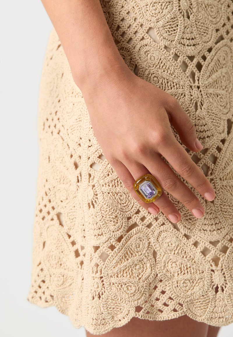 Yellow resin ring with an octagonal lavender stone, featuring clear accents, worn on a hand against a beige crocheted lace background.