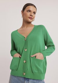 Zielony kardigan z dekoltem w serek, z przodu zapinany na guziki, z dwoma kieszeniami naszytymi. Wykonany z miękkiego, elastycznego materiału z prążkowanymi mankietami i dolnym brzegiem.
